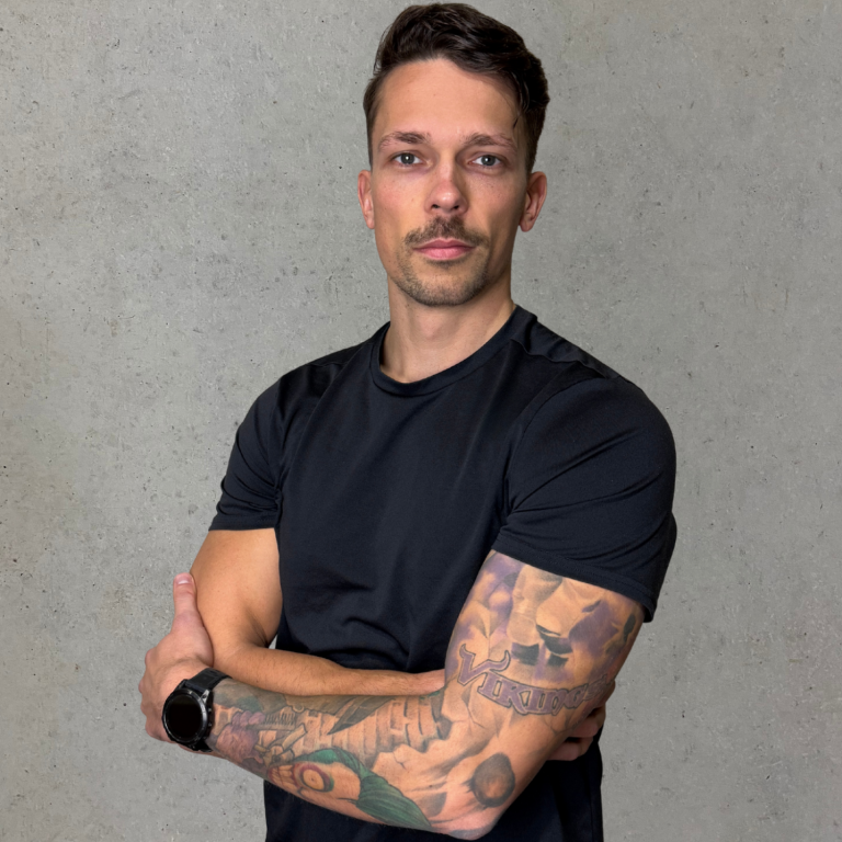 Vivi Junge Frau mit kurzem Haar, lächelt und steht mit verschränkten Armen vor grauem Hintergrund.